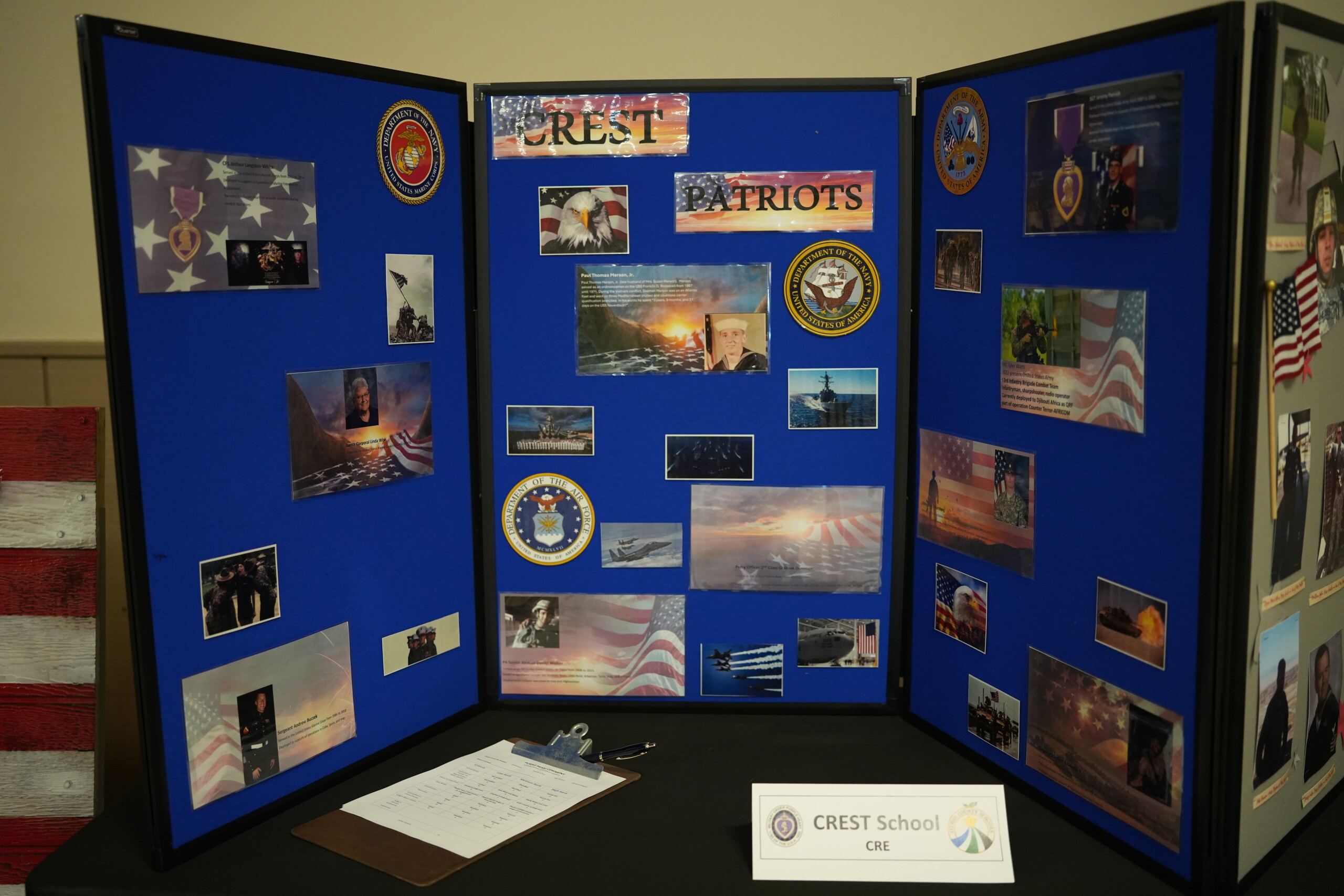 A three-panel blue display board features photos, American flags, and military branch seals, with a sign reading "CREST School" in front.